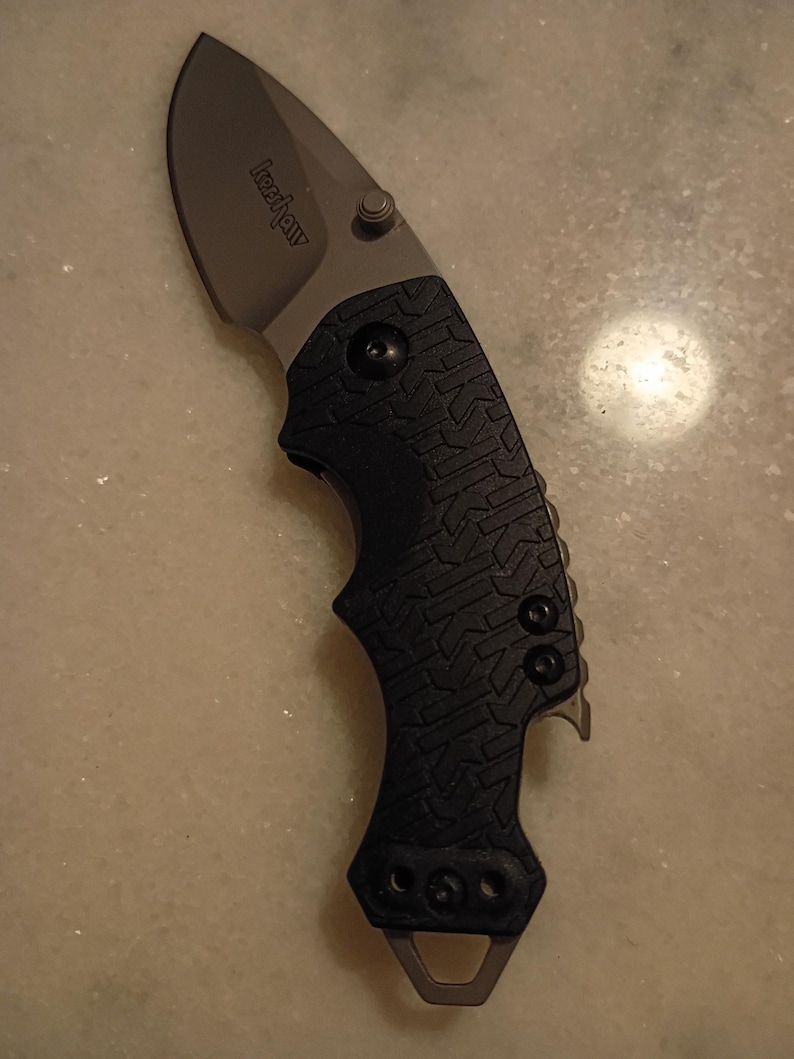 May include: A black and silver folding knife with a curved blade. The knife has a textured black handle and a silver blade with the brand name "Kershaw" printed on it. The knife also has a small metal loop at the base of the handle.