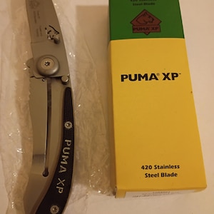May include: A silver folding knife with a black handle and the text "PUMA XP" is next to a yellow and green box. The box has the text "PUMA XP" and "420 Stainless Steel Blade". The knife is in a clear plastic wrapper.