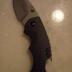 May include: A black and gray folding knife with the brand name "Kershaw" on the blade. The blade is curved and has a silver finish. The handle is black with a textured pattern and a lanyard hole.