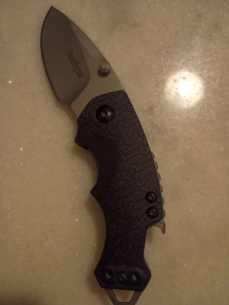 May include: A black and gray folding knife with the brand name "Kershaw" on the blade. The blade is curved and has a silver finish. The handle is black with a textured pattern and a lanyard hole.