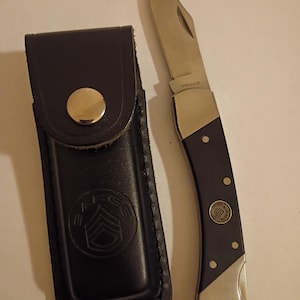 May include: A black folding knife with a silver blade and a dark blue handle, next to a black leather sheath. The knife handle has silver accents and rivets. The sheath has a gold snap closure and a military emblem.