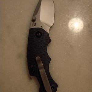 May include: A folding pocket knife with a silver blade and a black textured handle. The knife has a pocket clip and the brand name "Kershaw" is visible. The blade is curved and the handle has a secure grip design.