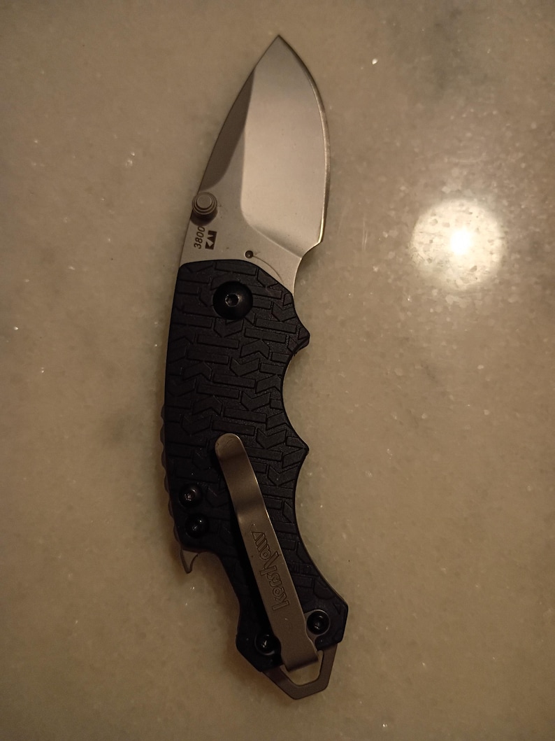May include: A folding pocket knife with a silver blade and a black textured handle. The knife has a pocket clip and the brand name "Kershaw" is visible. The blade is curved and the handle has a secure grip design.