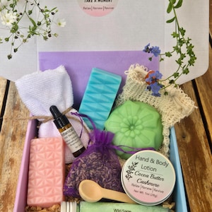 May include: A gift box containing a pink geometric soap, a blue geometric soap, a green flower-shaped soap, a white washcloth, a small bottle of oil, a purple organza bag of dried lavender, a wooden spoon, and a tin of cocoa butter lotion.