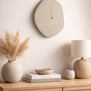 Pode incluir: Um relógio de parede bege em forma de seixo com ponteiros dourados está pendurado na parede. Abaixo, um armário de madeira exibe um vaso de capim-dos-pampas seco, livros, uma pequena tigela de madeira, um castiçal de cerâmica e uma lâmpada canelada.