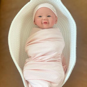 May include: A realistic baby doll with a smiling expression, wearing a light pink ribbed swaddle and a matching light pink ribbed beanie, resting inside a white woven basket with a textured, rope-like construction and an oval shape.