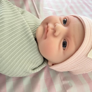 May include: A baby wrapped in a light green ribbed blanket, wearing a pink ribbed knit hat with a small label that reads "Little Hearts". The baby is lying on a pink and white checkered surface.