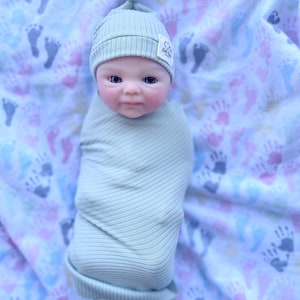 May include: A baby doll wrapped in a light green ribbed blanket and matching beanie. The beanie has a small tag with a heart and text. The doll is lying on a blanket with colorful handprints.