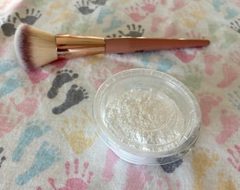 Könnte beinhalten: Ein Make-up-Pinsel mit einer roségoldenen Zwinge und einem beigen Griff, platziert neben einem kleinen, runden, durchsichtigen Kunststoffbehälter mit feinem weißen Puder. Der Pinsel hat weiche, dichte Borsten, die von Weiß an den Spitzen zu einem dunklen Rotbraun an der Basis übergehen.