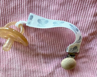 May include: A translucent amber-colored pacifier attached to a white fabric strap featuring a repeating gray elephant pattern. The strap is secured with a metal clip and a round, light-colored wooden bead at the end of the attachment.