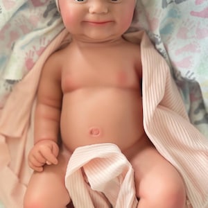 May include: A realistic baby doll with light skin, blue eyes, and a soft, rounded face. The doll wears a pale pink ribbed beanie and is partially wrapped in a matching pale pink ribbed fabric, showcasing detailed facial features and a navel.