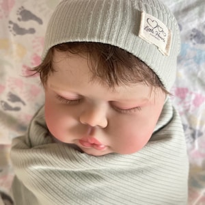 May include: A lifelike baby doll wearing a light green ribbed knit cap and swaddled in a matching blanket. The cap has a small tag that reads "Little Hearts Nursery". The doll has closed eyes and a peaceful expression.