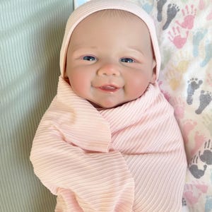 May include: A lifelike baby doll with blue eyes and a smiling expression, wrapped in a light pink ribbed blanket and wearing a matching cap. The doll is lying on a patterned blanket with colorful hand and footprint designs.