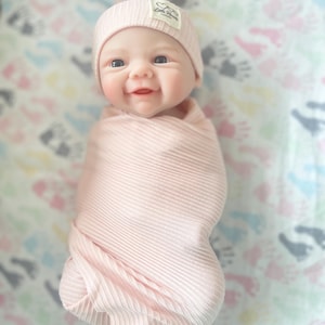 May include: A baby doll wrapped in a light pink ribbed knit blanket and matching beanie. The beanie has a small tag that reads "Little Hearts". The doll has a smiling expression and is lying on a patterned sheet with colorful footprints.