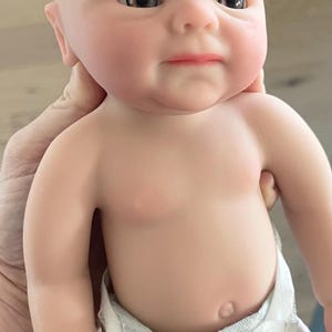 May include: A realistic baby doll with a smooth, light-toned complexion, dark brown eyes, and painted eyebrows. The doll has a bald head, a defined belly button, and is wearing a white disposable diaper with a textured, paper-like appearance.
