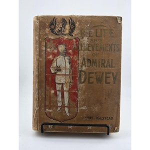 Puede incluir: Libro antiguo titulado "The Life and Achievements of Admiral Dewey" de Murat Halstead. La portada es marrón con letras doradas y una ilustración roja y blanca de un hombre uniformado. El libro está sujeto por un soporte de metal negro.