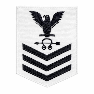 May include: White rectangular patch featuring a black eagle with outstretched wings above a symbol and three chevrons. The patch is likely an insignia or emblem, possibly for a military or uniformed service.