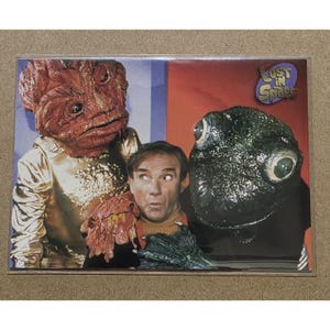 May include: A trading card featuring characters from the television show "Lost in Space." The card shows three figures: two aliens and a man. The aliens have large heads and appear to be wearing costumes. The man is in the center, looking surprised.