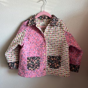 May include: A quilted jacket with a floral pattern in pink, white, and black. The jacket has a collared neckline, two pockets, and gold-colored buttons. The label reads "Dear Vanessa".