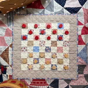 May include: A square fabric game board featuring a patchwork grid of floral, solid, and gingham squares. The board is bordered by a brown and white gingham trim. Red and light wood circular game pieces are arranged on the quilted surface.