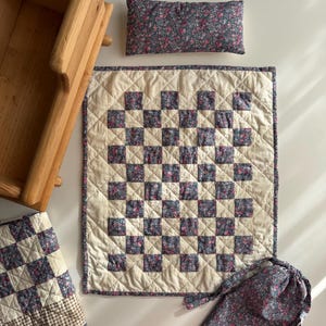 May include: A wooden cradle, a rectangular pillow, and a quilted blanket featuring a checkerboard pattern of cream fabric and dark blue floral fabric. A matching drawstring bag with a small rectangular label is included in the set.
