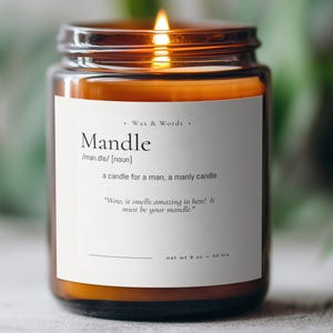 May include: A lit candle in a brown glass jar with a white label. The label reads "Mandle" with a definition and a quote. The candle is described as a candle for a man, with a net weight of 9 oz and a burn time of 50 hours.