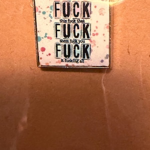 May include: A square pin badge featuring the word "FUCK" repeated three times in a bold font. The badge has a white background with colourful paint splatters and the phrases "this fuck that", "them fuck you", and "it fucking all".