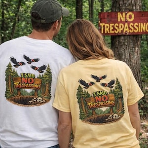 May include: Two people wearing white and yellow t-shirts with a graphic design on the back. The design features a sign that reads "I Said No Trespassing" with a skull, birds, and trees. A "No Trespassing" sign is visible in the background.