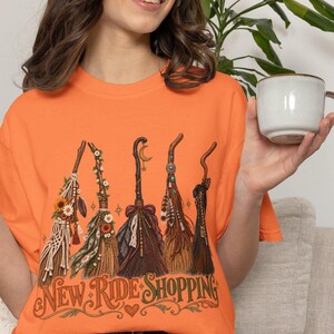 May include: An orange t-shirt featuring a graphic of five decorated broomsticks with flowers, feathers, and lace. Below the broomsticks, the text reads "NEW RIDE SHOPPING" in a stylized font, accompanied by a small heart and decorative flourishes.