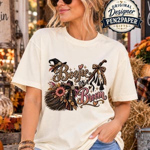 May include: An ivory short-sleeve t-shirt featuring a graphic print of a witch hat, a broomstick, and the text "Boujee ON A Broom". The design includes leopard print patterns, a sunflower, and decorative bows in shades of brown, pink, and black.
