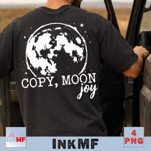 Puede incluir: Camiseta negra con un gráfico blanco de una luna llena y estrellas. El texto "COPY, MOON joy" está debajo del diseño de la luna. La camiseta es de color oscuro y parece estar hecha de un material suave.