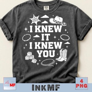 May include: A charcoal gray short-sleeve t-shirt featuring white graphic prints including a sheriff badge with the text TO INFINITY AND BEYOND, a cowboy hat, cowboy boots, a lasso, alphabet blocks, and the text I KNEW IT I KNEW YOU.