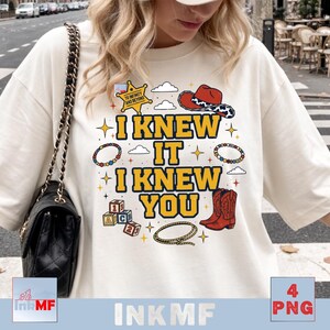 May include: A cream-colored t-shirt featuring a graphic print with the text I KNEW IT I KNEW YOU in yellow letters. The design includes a sheriff badge, a red cowboy hat, a lasso, alphabet blocks, beaded bracelets, and small stars.
