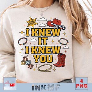 May include: A beige crewneck sweatshirt featuring a graphic print with the text I KNEW IT I KNEW YOU in yellow with blue outlines. The design includes a yellow sheriff star, a red cowboy hat, alphabet blocks, a lasso, and colorful beaded bracelets.