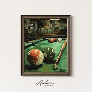 May include: A framed print of a pool table scene. The artwork features a green felt table with several billiard balls, including a striped orange and white ball and a solid green ball with the number 8. A pool cue rests on the table.