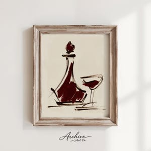 Puede incluir: Obra de arte enmarcada que representa una jarra de vino y una copa de vino. La jarra es de color burdeos intenso con una parte superior redondeada, y la copa de vino está parcialmente llena. La obra de arte está en un marco de madera envejecida.
