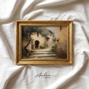May include: A framed landscape painting depicts a European village scene with stone steps, buildings, and a tree. The artwork is in a gold-colored frame. The painting has a vintage aesthetic.