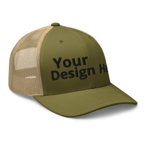 May include: An olive green trucker hat with a tan mesh back and a solid front panel. The front panel features the black embroidered text Your Design Here. The hat has a structured crown, a curved brim, and a button on the top center.