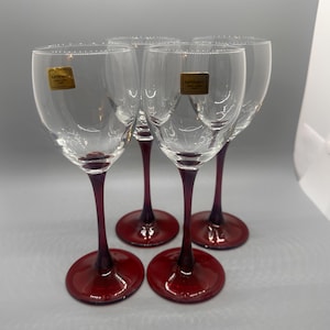 May include: Four clear wine glasses with burgundy stems and bases. Each glass has a gold label near the rim. The stems and bases are a deep red color, and the bowls are transparent.