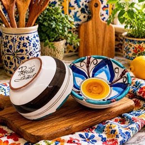 May include: A white ceramic mug with a black handle and the text "But First, Coffee" in a bold, black font. The mug is set against a blurred background, suggesting a cozy setting.