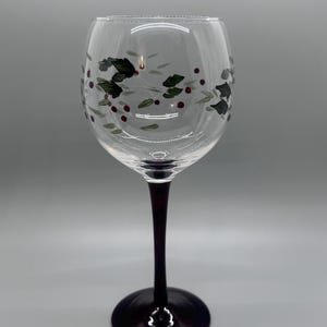 May include: A clear wine glass with a dark purple stem and base. The glass is decorated with green leaves and red berries. The design is painted on the glass, creating a festive look. Perfect for serving wine or other beverages.