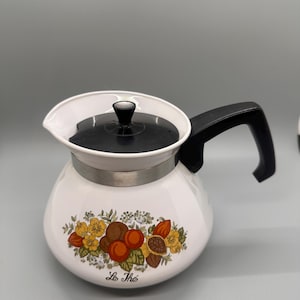 May include: White ceramic teapot with a black handle and lid. The teapot features a colorful floral design with the text "Le Thé" in a script font. The design includes orange, yellow, and brown flowers and fruits.
