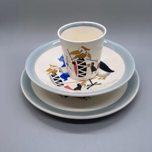 May include: A vintage children's dish set featuring a white cup and two plates. The set has a light blue rim and is decorated with cartoon owls and other whimsical figures in red, black, and blue. The set is designed for children.