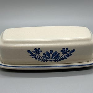 May include: Cream-colored ceramic butter dish with a lid, featuring a blue floral design. The dish has a rectangular shape with rounded edges and a matching base with a blue border. A classic kitchenware item.