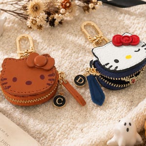 May include: Two round zippered pouches shaped like cat faces with bows. One is brown with a letter C charm, and the other is white with a red bow, blue base, and letter D charm. Both feature gold-tone metal clasps and zipper pulls.