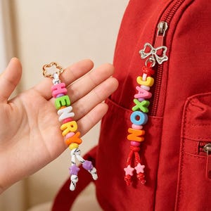 Op de afbeelding: Twee gepersonaliseerde rits-hangers met kleurrijke plastic letterkralen die de namen HENRY en JAXON vormen. De bedels zijn geregen aan wit en rood koord met kleine stervormige kralen aan de uiteinden en bevestigd aan zilverkleurige metalen sluitingen.