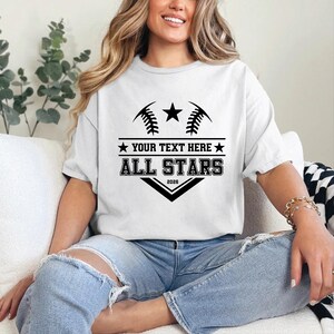 May include: White t-shirt with a black graphic design featuring baseball stitching, a star, and the text "YOUR TEXT HERE" above "ALL STARS 2026". The shirt is worn by a person.