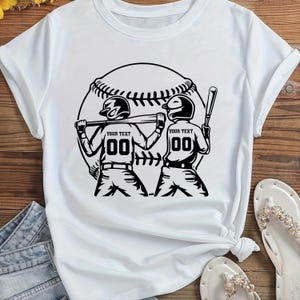 May include: White t-shirt with a black graphic of two baseball players with helmets and bats, set against a baseball. The text "YOUR TEXT" and the number "00" are on the players' uniforms. The shirt is on a wooden surface.