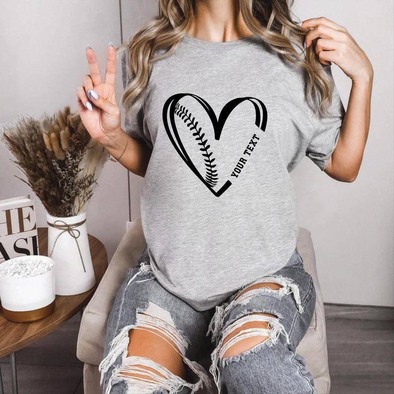 May include: A light grey t-shirt with a black heart design incorporating baseball stitching on the left. The right side of the heart has the text "YOUR TEXT". The shirt is made of a soft material.