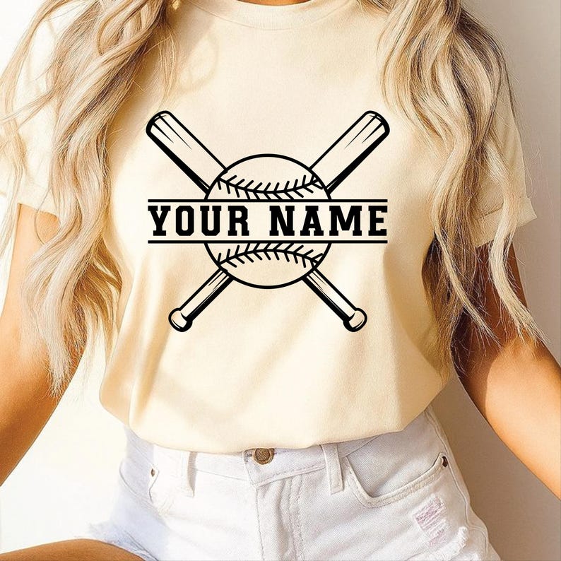 May include: A cream-coloured t-shirt with a baseball design. The design includes crossed bats, a baseball, and the text "YOUR NAME" in black. The shirt is made of a soft material. The design is centered on the front of the shirt.